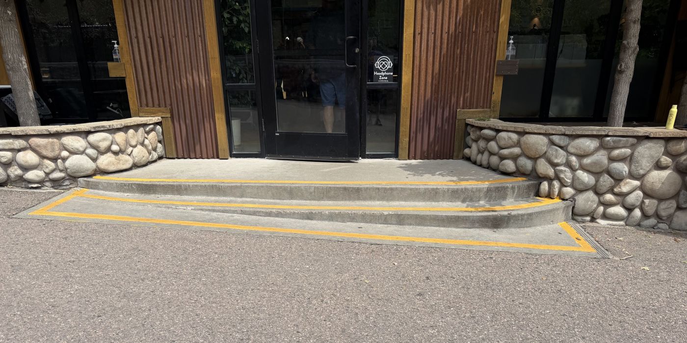 Two concrete steps with yellow nosings at a building entrance. The bottom step lands on a sloped concrete wedge that runs down to the pavement.